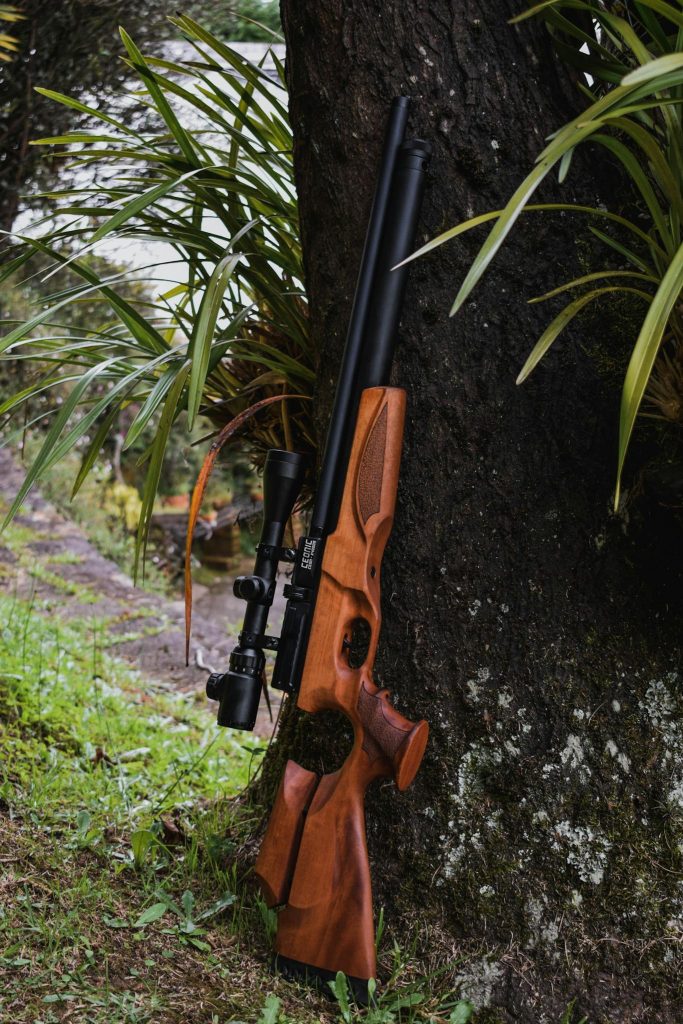 A detailed view of a wooden rifle set against a tree in a lush outdoor setting.